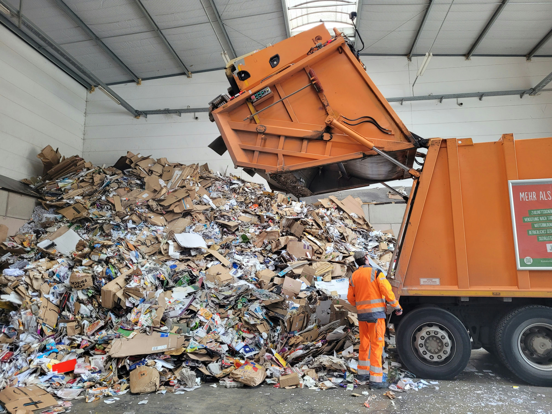 A worker at a recycling facility standing beside a garbage truck and a massive pile of cardboard waste